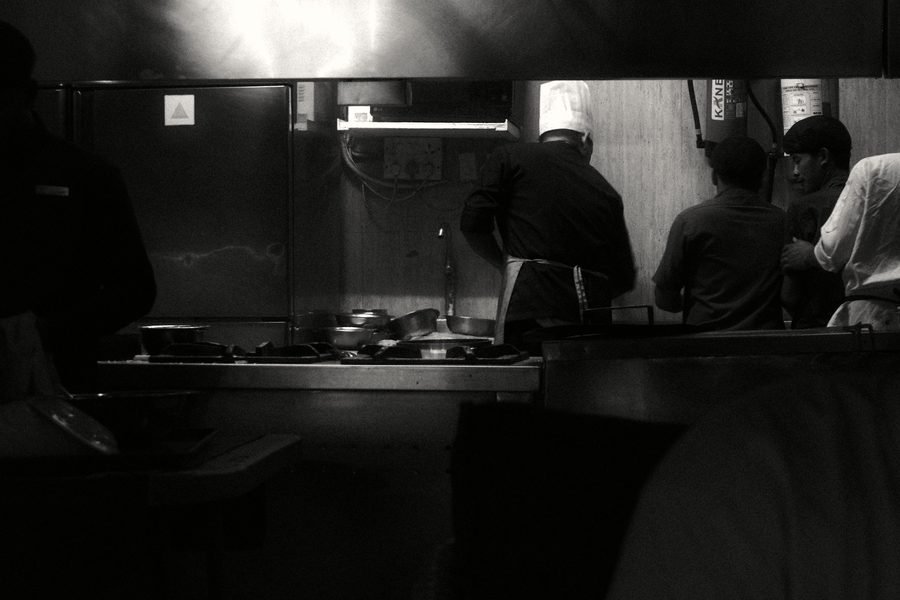 Kitchen staff working at a line in a dim commercial kitchen, seen from behind.