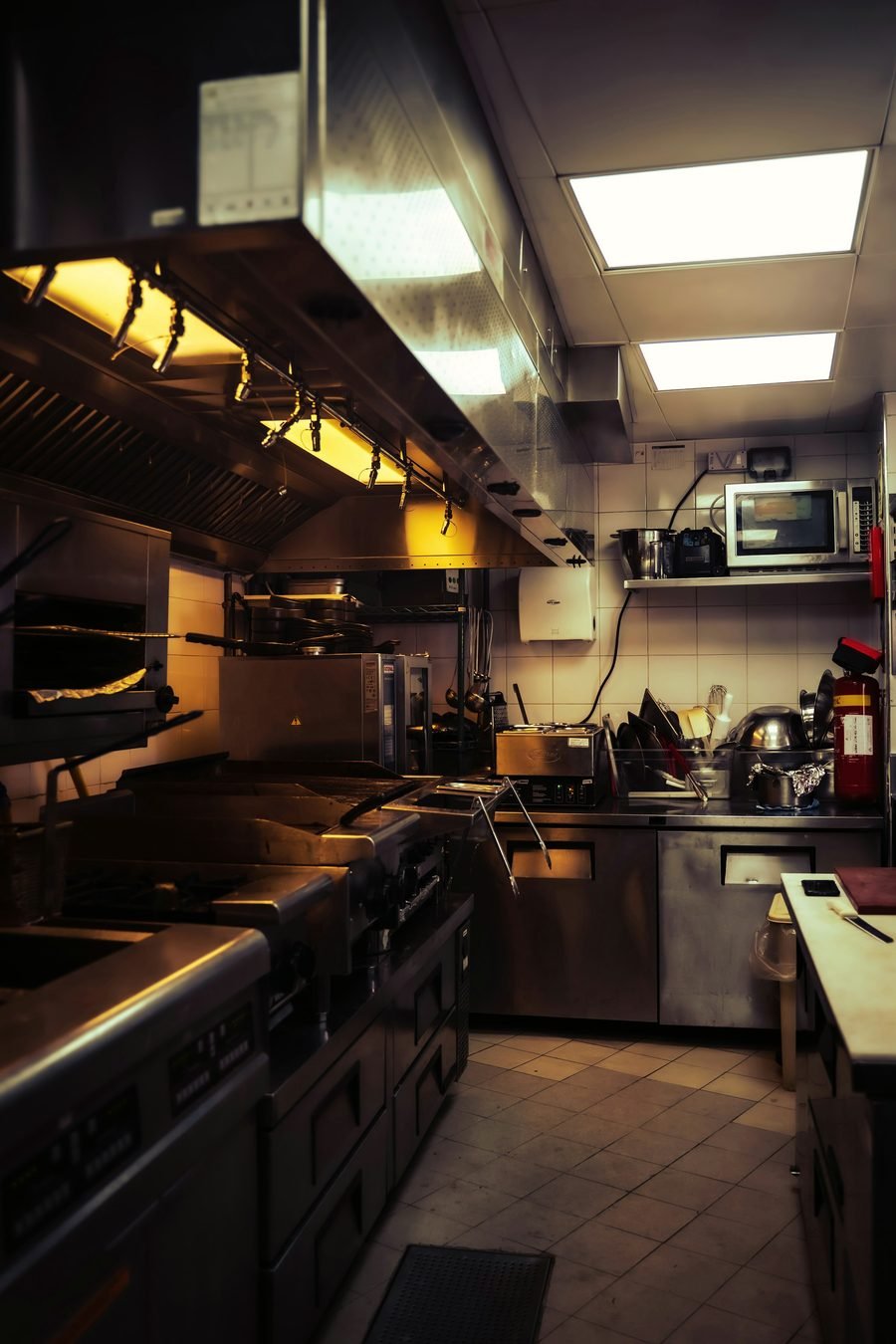 A tight commercial kitchen line lit warm, with steel prep tables and a fire extinguisher on the wall.