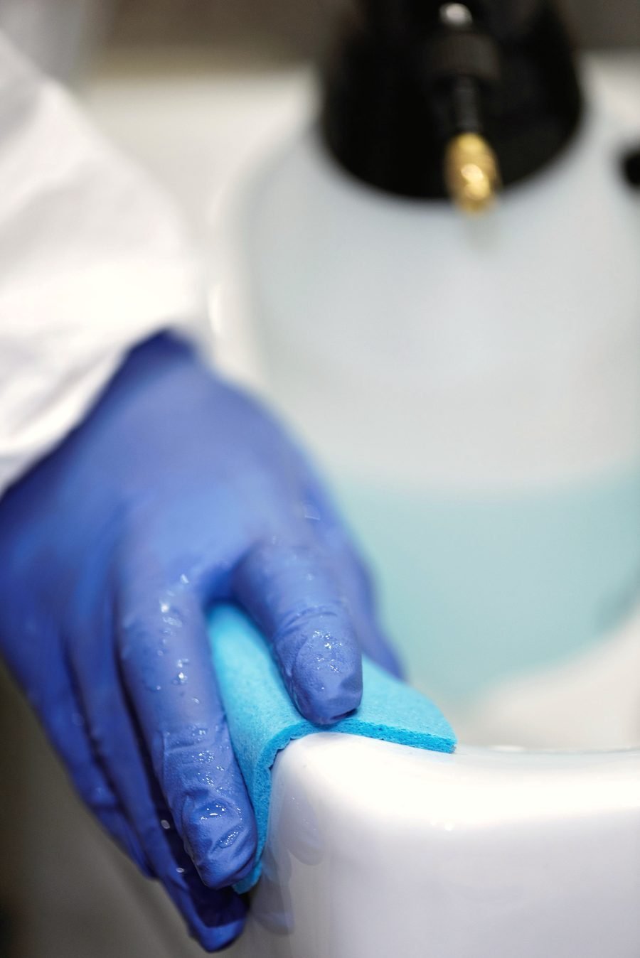 A gloved hand cleaning a stainless steel sink basin with a sponge.